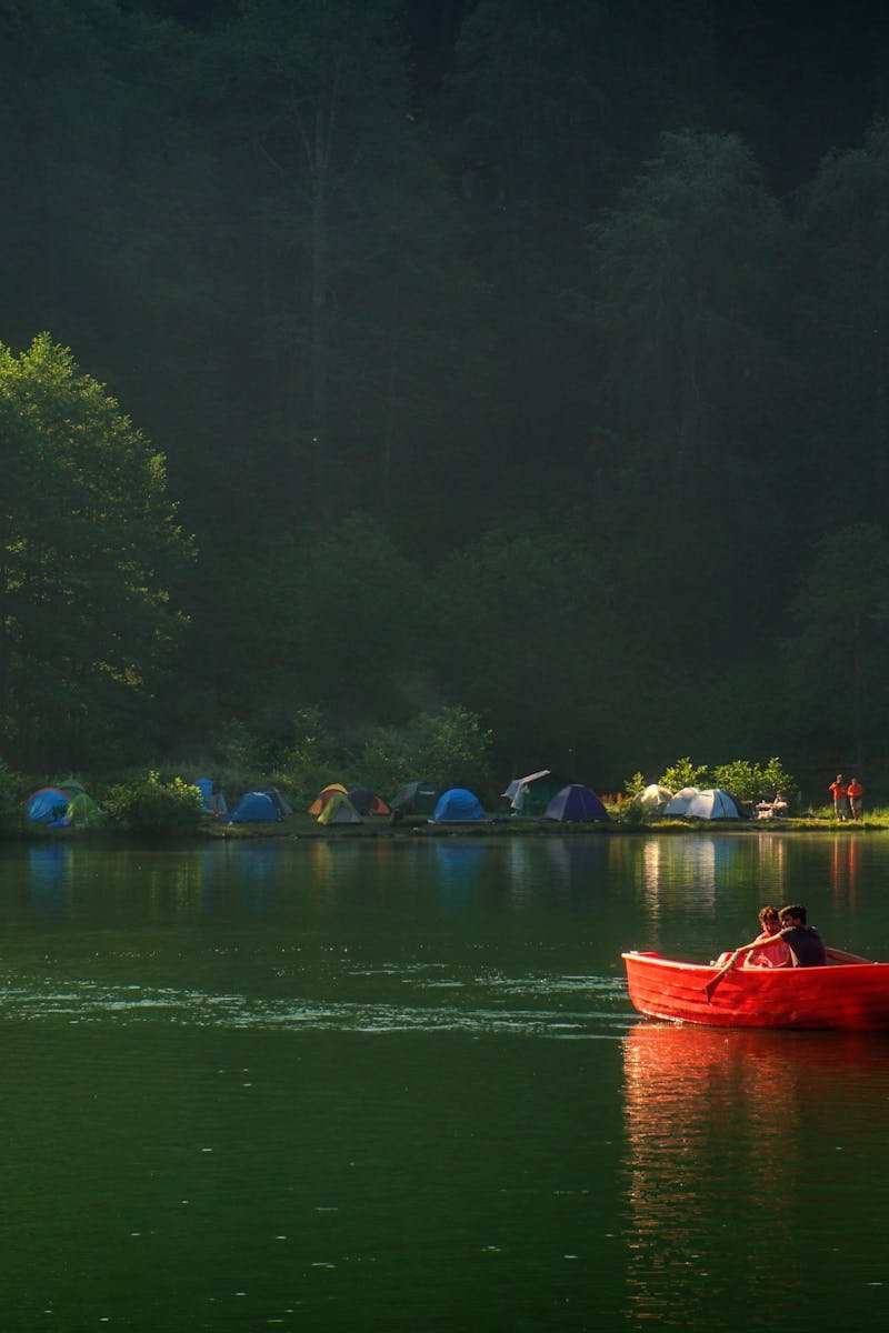 Two people in a red boat on a serene lake near a forest campsite.
